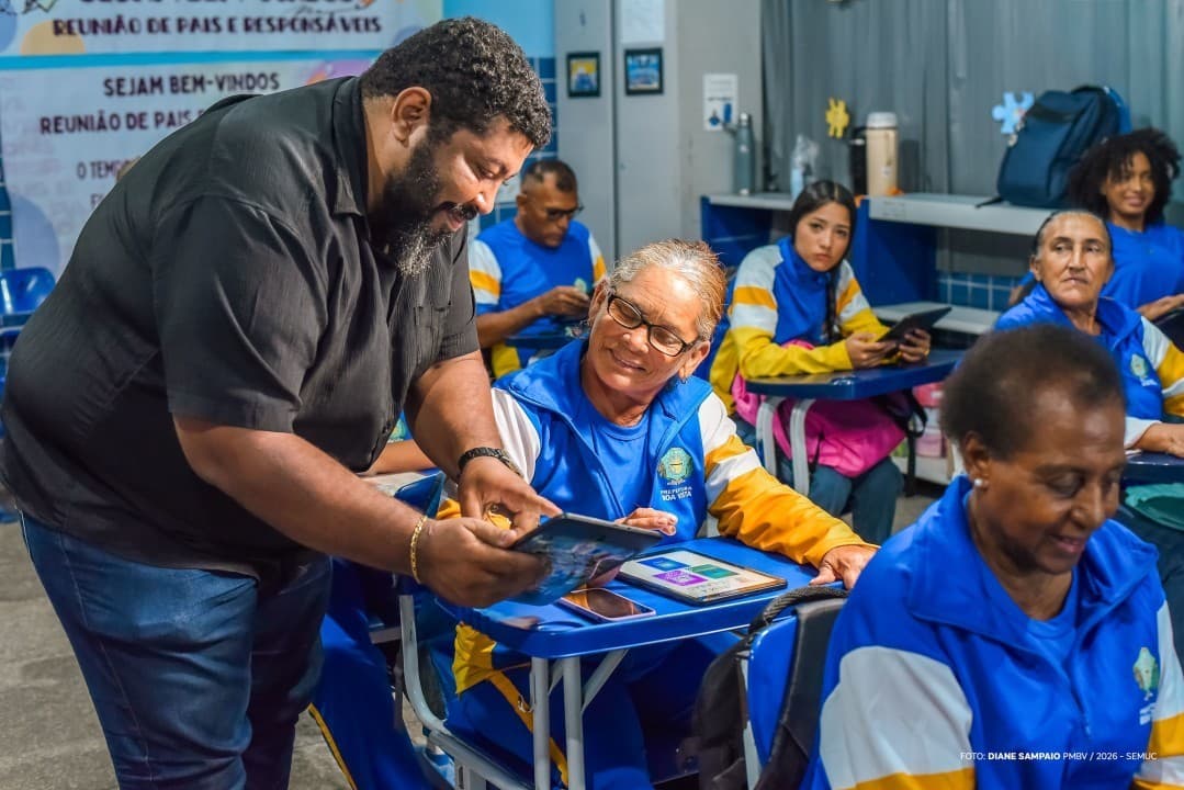Mãos de idosa seguram tablet dentro de sala de aula iluminada.