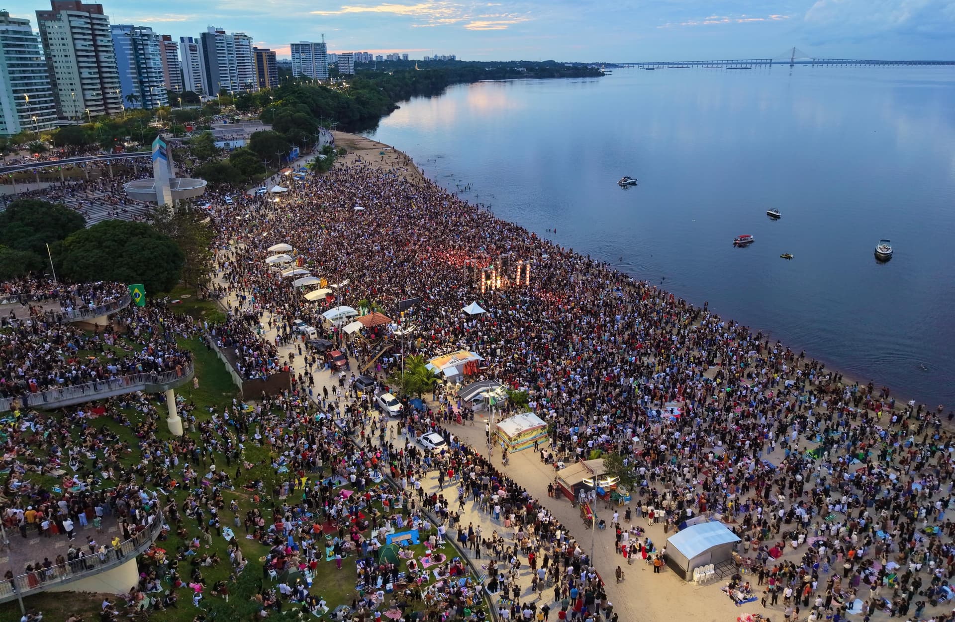 Multidão ocupa a areia da Praia da Ponta Negra ao amanhecer, com luzes de palco ao fundo.
