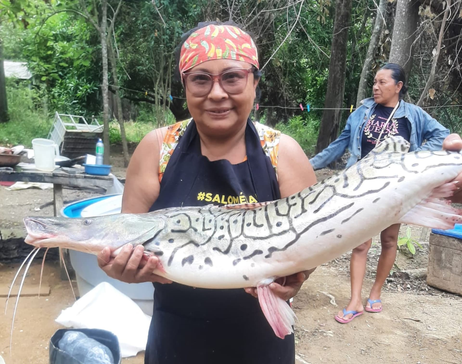Dona Rosinha serve prato de peixe frito em mesa sob mangueiras.