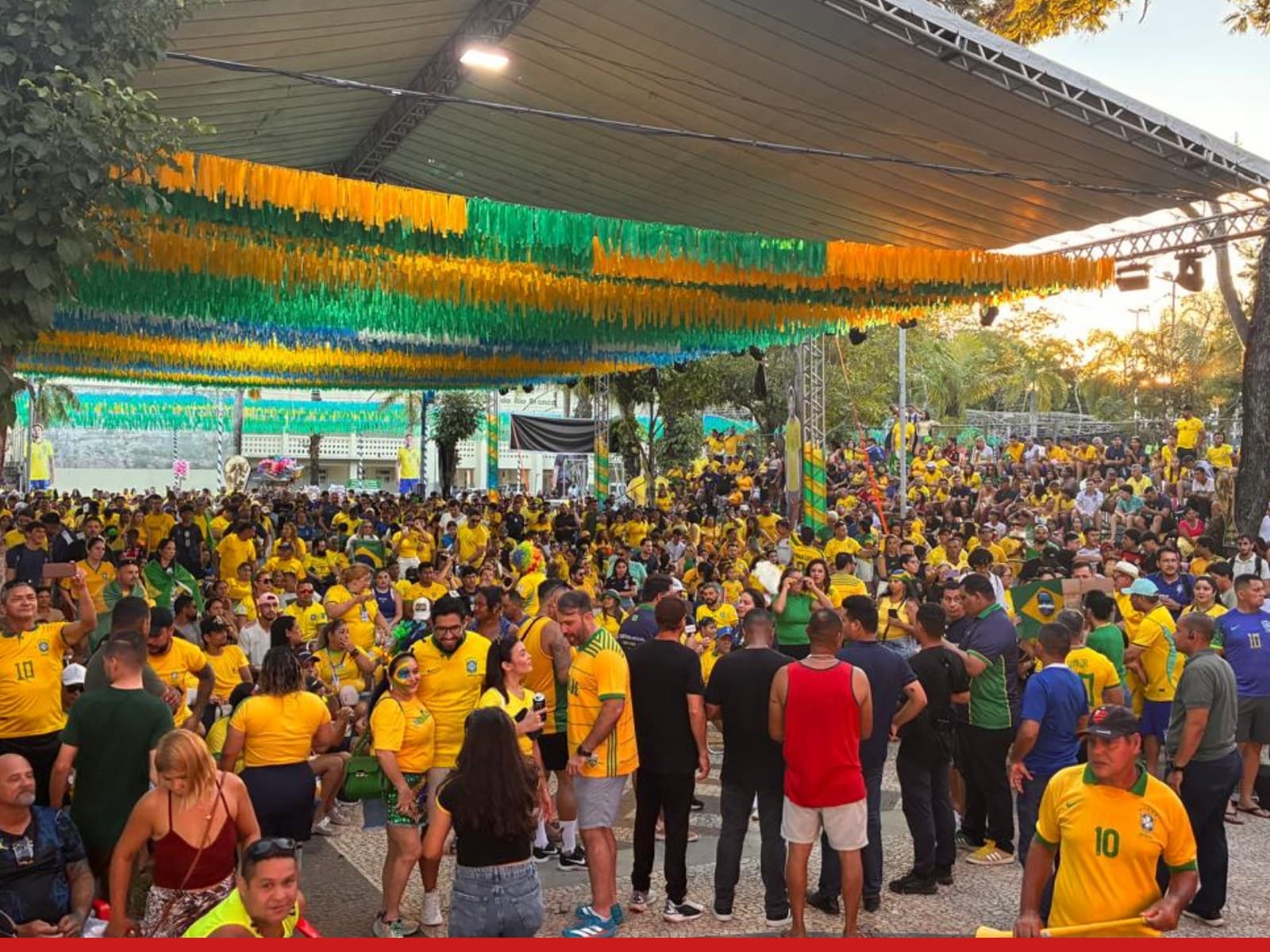 Torcedores vestidos de verde e amarelo se aglomeram em frente ao telão na Praça da Revolução ao entardecer.