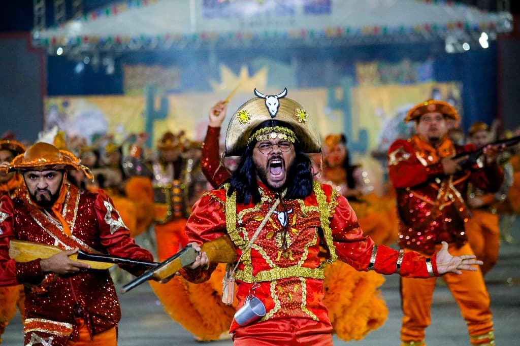 Brincantes de boi-bumbá com fantasias coloridas dançam em arena iluminada durante festa junina noturna.