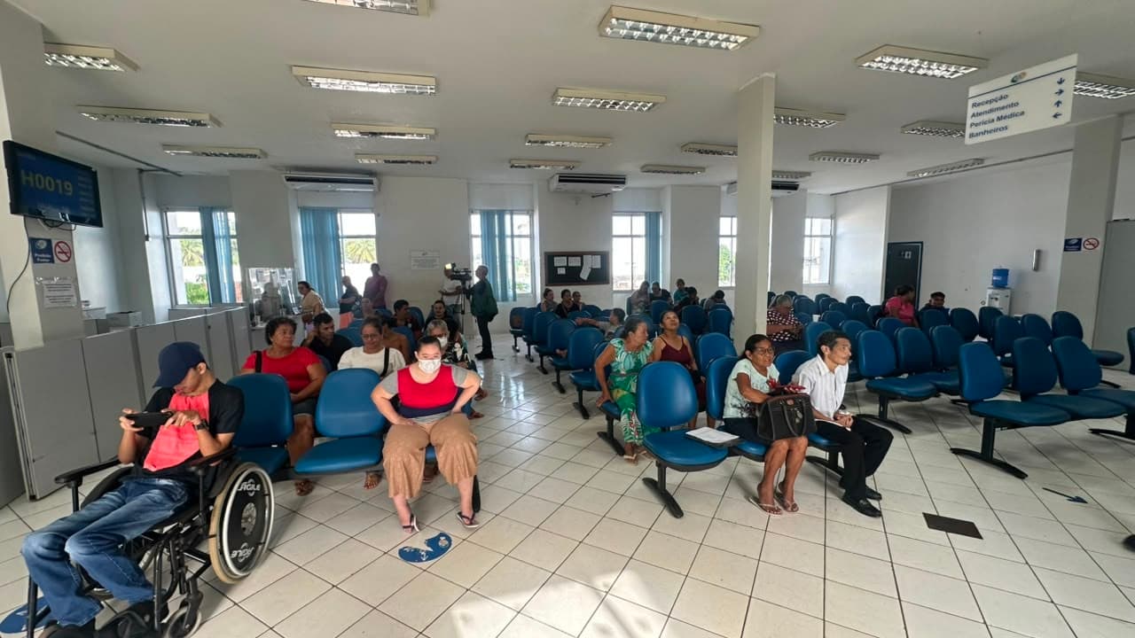 Fila de pessoas aguarda atendimento sentada em cadeiras plásticas na calçada da agência do INSS em Rio Branco.