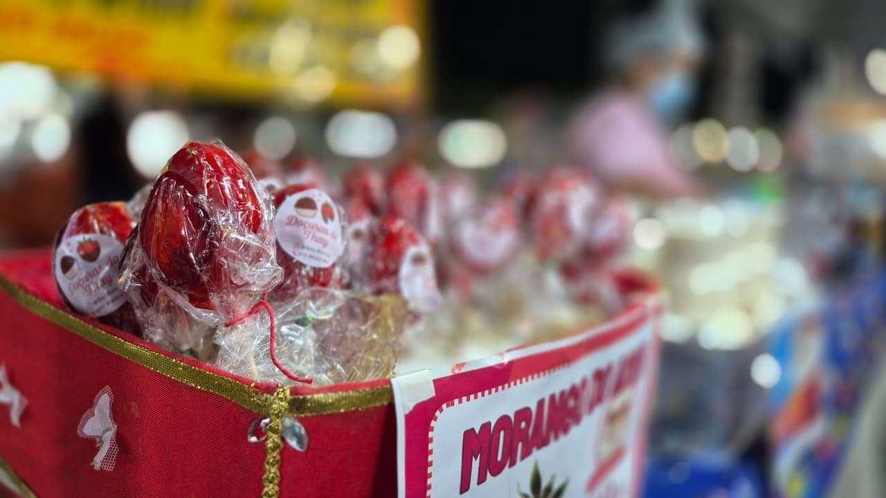 Mãos seguram morangos cobertos por chocolate e granola em ambiente iluminado de festa junina em Boa Vista.