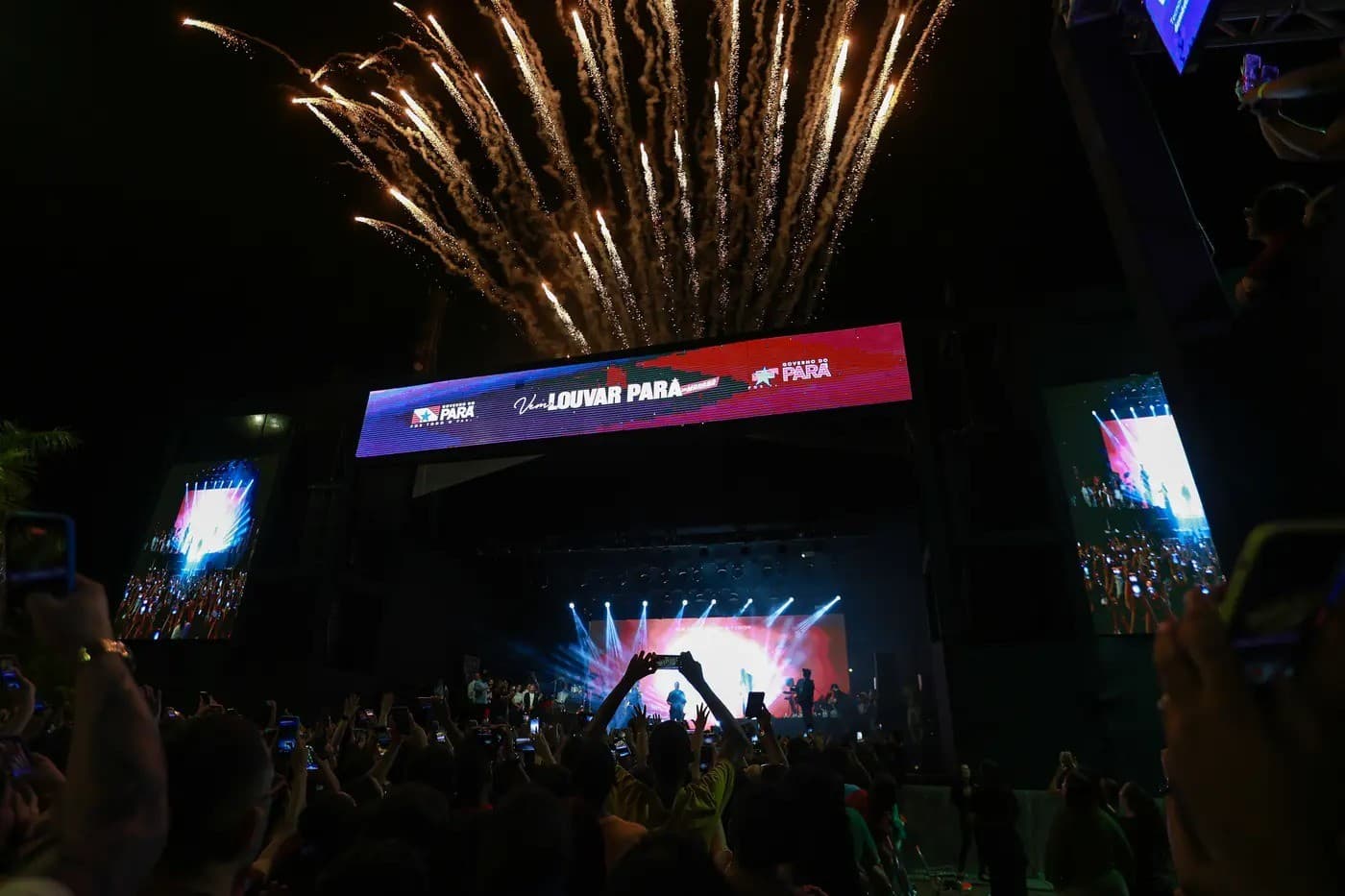 Palco iluminado ao entardecer em estacionamento de estádio, com público em cadeiras de plástico ao fundo.
