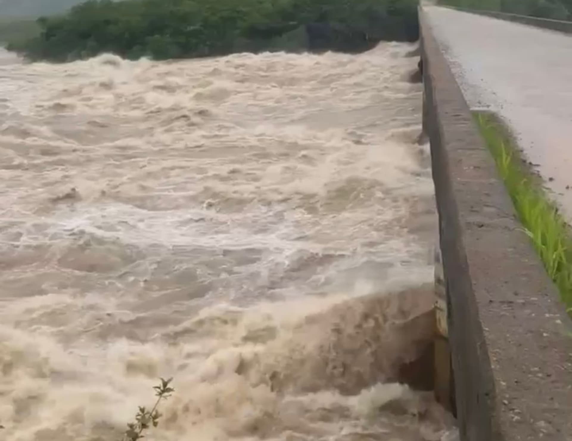 Vista de estrada alagada às margens de rio em Uiramutã, com correnteza forte e vegetação submersa.