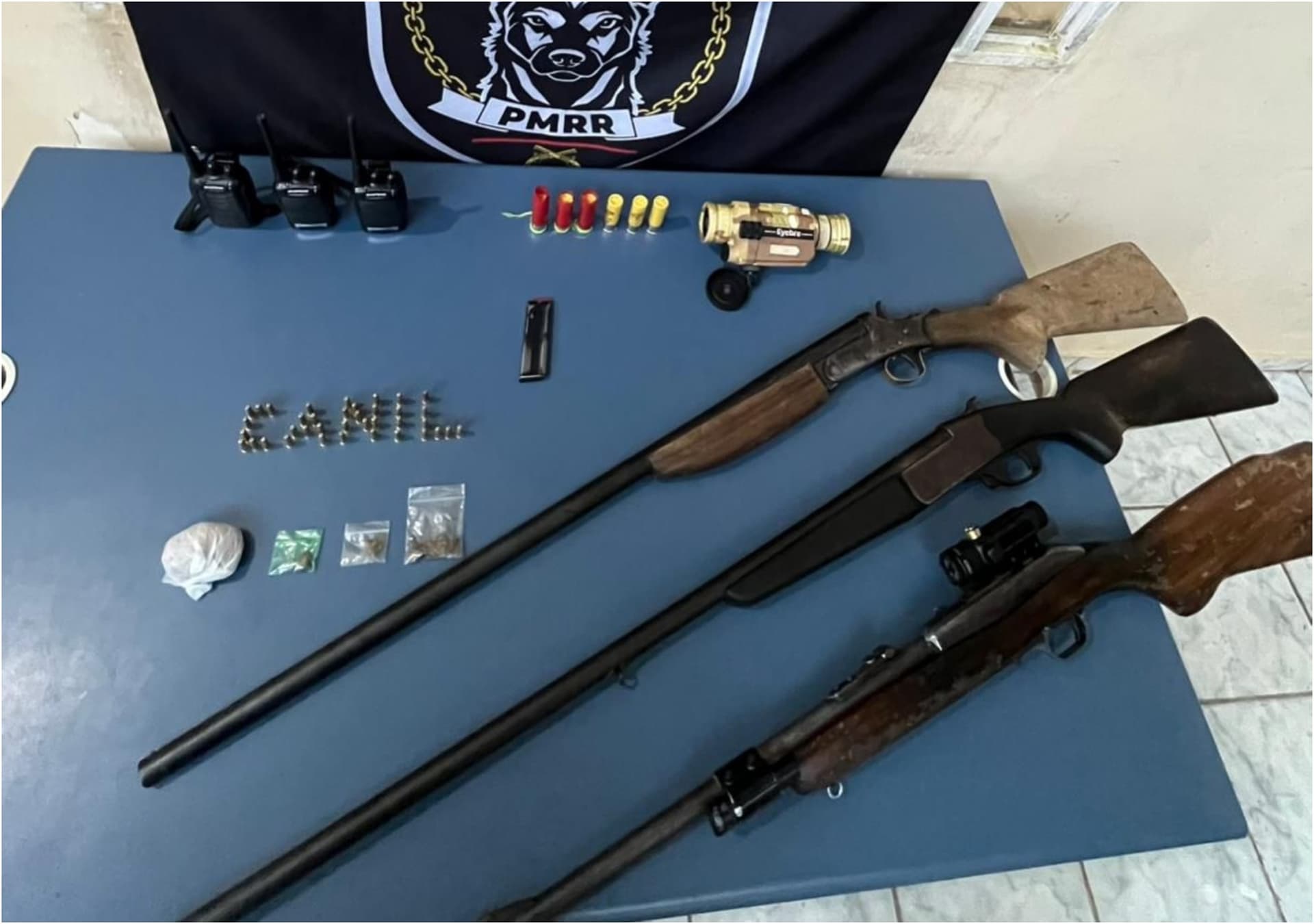 Mochila preta aberta sobre o asfalto com armas de fogo, rádios e munições expostos ao lado.