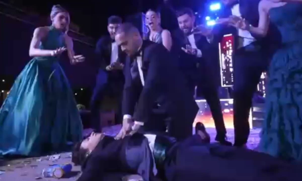 Jovens vestidos de formandos fazem coreografia de massagem cardíaca em palco iluminado.