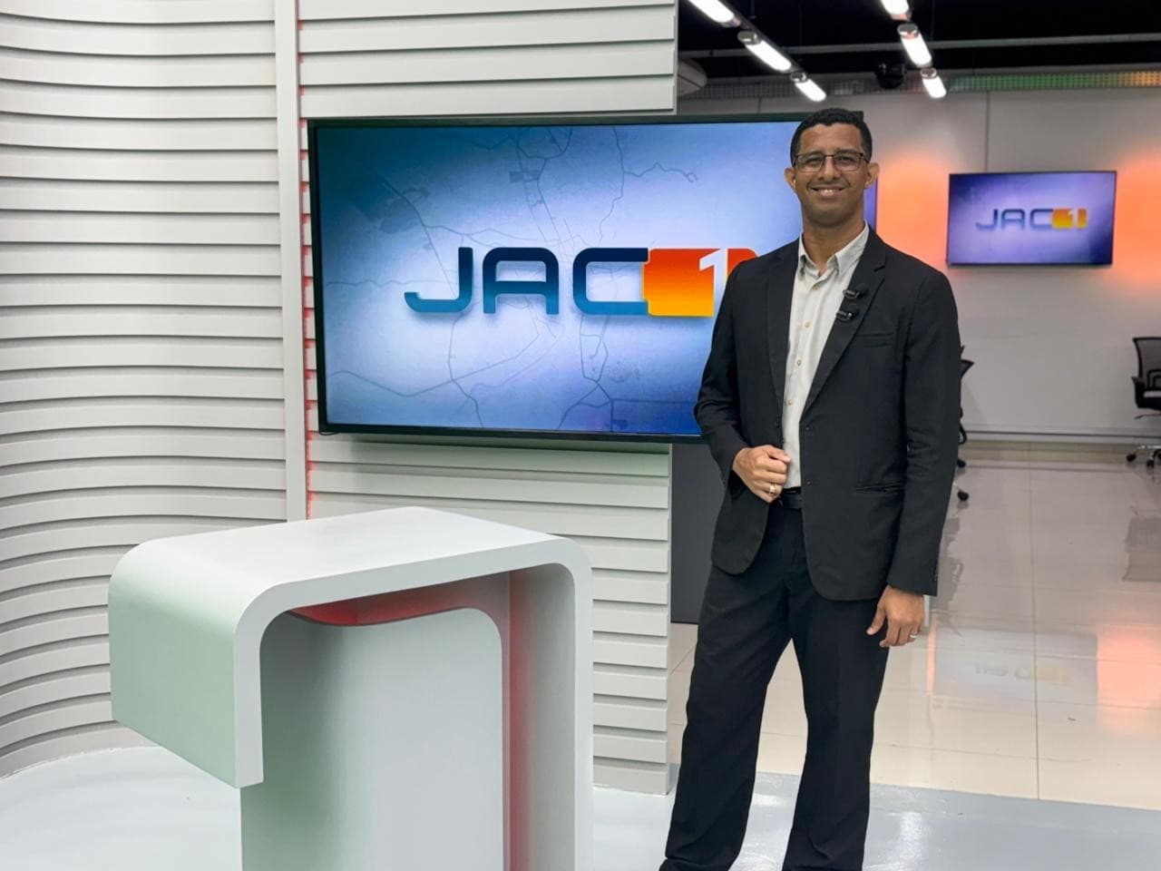 Murilo Lima em estúdio de televisão durante gravação do JAC1, com microfone e papéis à frente.