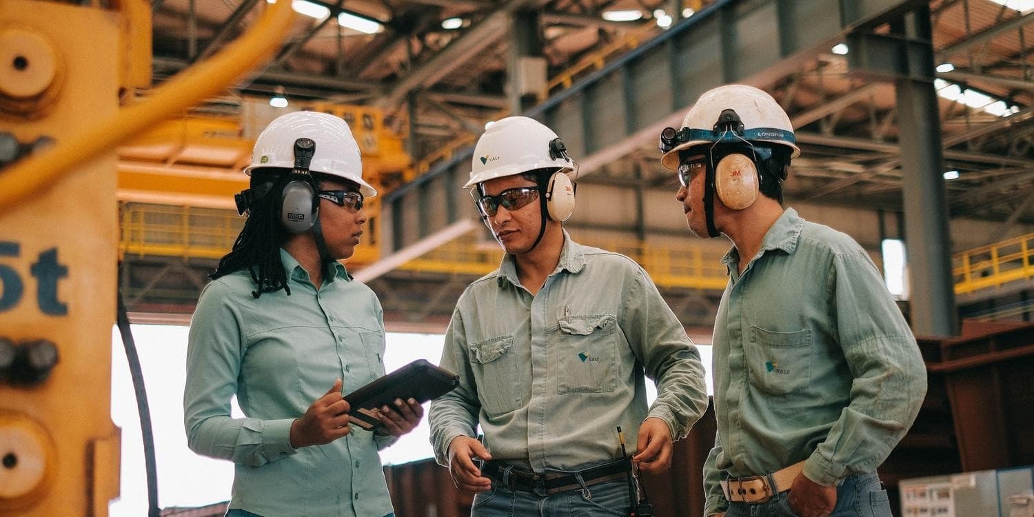 Planta industrial de mineração ao fundo com operadores de equipamentos em primeiro plano.