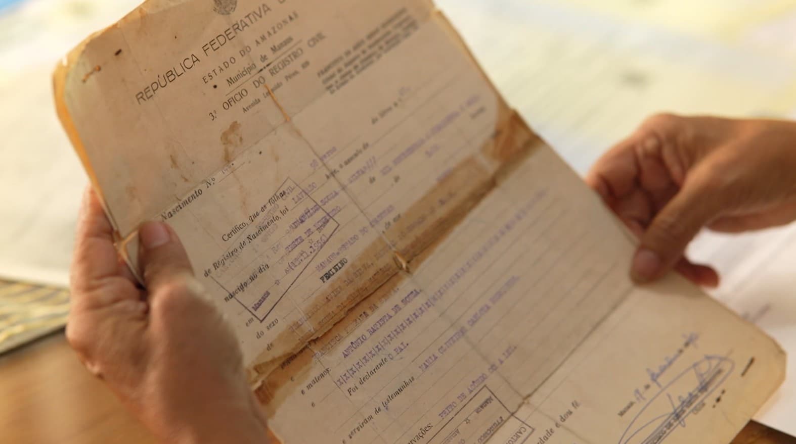Mãos seguram certidões de nascimento e carteiras de identidade sobre mesa de atendimento em cartório.