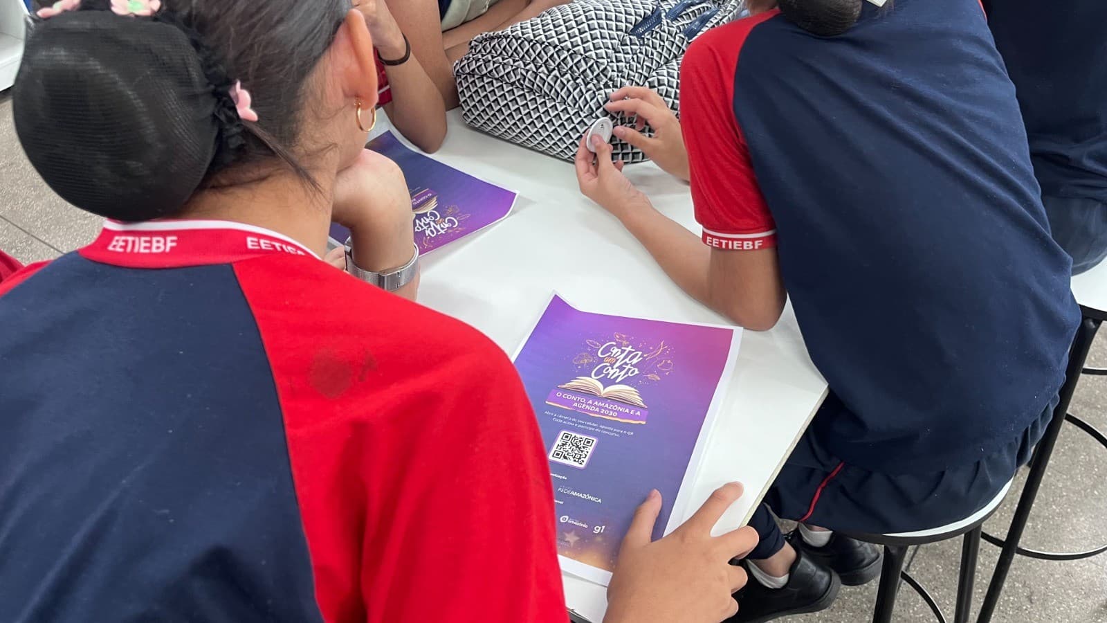 Estudante escreve em caderno com caneta em sala de aula iluminada pelo sol da manhã.