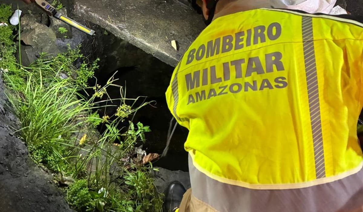 Boca de bueiro em rua asfaltada de Manaus à noite, com viatura estacionada ao fundo.